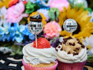 Colorful cupcakes decorated with whipped cream, strawberries and disco ball lollipops against a floral background.