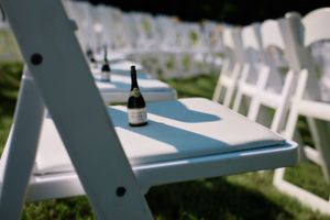 A single wine bottle sits on a white folding chair at an outdoor wedding setup.