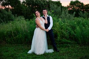 Couple in wedding attire poses together in a lush green field at sunset.