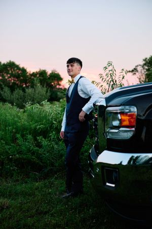 A suited person stands beside a black truck during sunset in a rural setting.