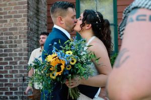 Wedding couple embracing with vibrant sunflower bouquet against brick wall.
