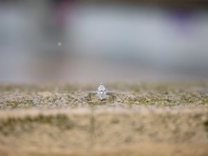 A diamond engagement ring sparkles while sitting on a textured sandy surface with soft bokeh in the background.