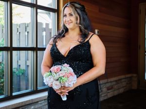 A person wearing a black sequined dress holds a pastel bouquet while standing by a window.