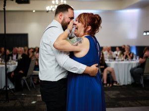 A couple share an intimate dance moment in formal attire at a wedding reception with guests seated in the background.