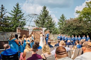 Outdoor wedding ceremony with blue-clad wedding party under metal gazebo arch surrounded by evergreen trees.