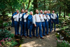 Groomsmen in navy suits and suspenders pose together on a garden path for a wedding photo.