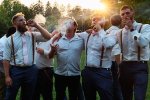 Groomsmen drinking and celebrating outdoors at sunset while wearing suspenders and dress shirts.