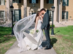 A couple shares a romantic moment outdoors as the bride's long veil flows in the wind at a stately venue.