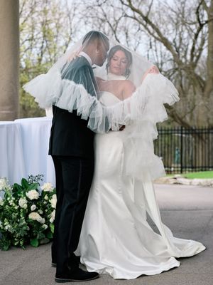 A bride and groom share an intimate moment outdoors with flowing veil and white floral decorations during their wedding ceremony.