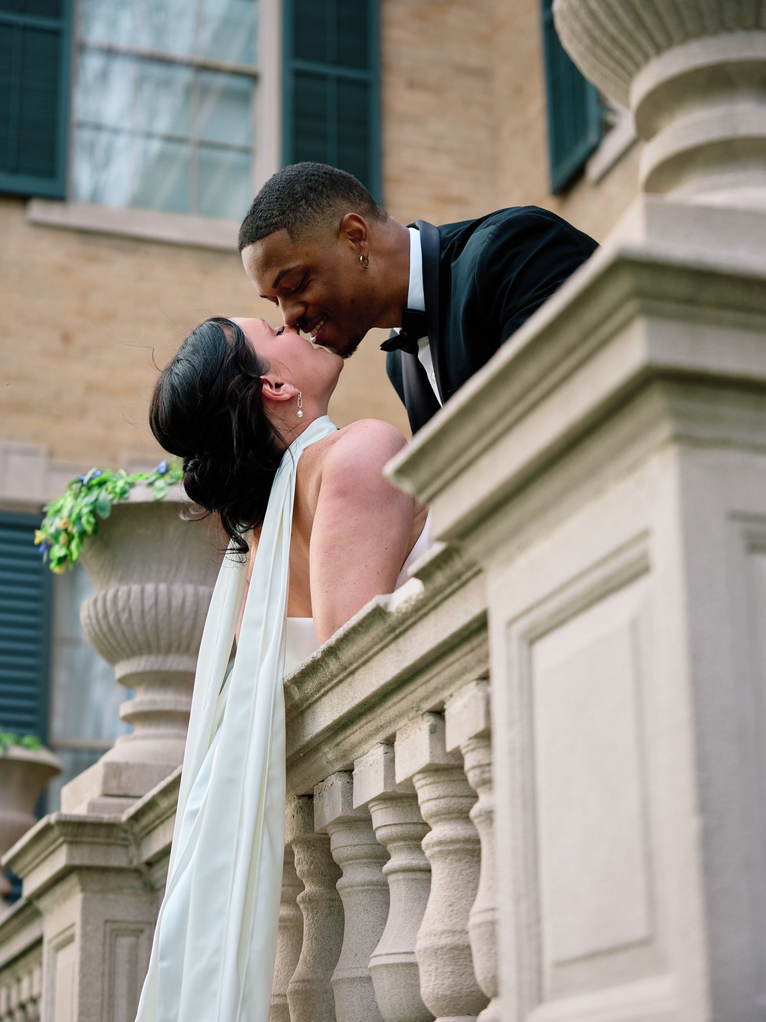 Newlyweds share a romantic kiss on an elegant stone balcony with classic architectural columns and green shutters.