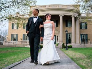 A newlywed couple walks along a path in front of a stately historic mansion with columns on a sunny spring day.