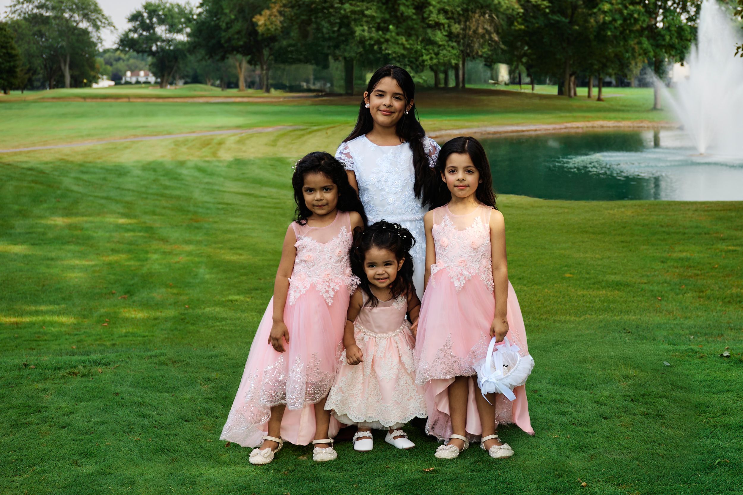 A group in matching pink dresses poses on a lush green lawn with a fountain in the background.