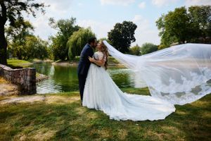A flowing white wedding dress with long veil caught in the breeze beside a peaceful pond in a garden setting.