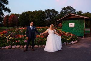 Wedding couple walks along a garden path lined with colorful flowers at dusk outside a rustic green building.