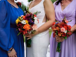 Wedding party members wearing blue, white and lavender dresses hold vibrant autumn colored floral bouquets.