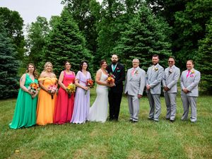 Wedding party in vibrant rainbow colored dresses and gray suits pose on grassy lawn with evergreen trees in background.