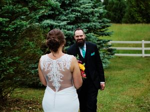 A smiling groom wearing a black suit gazes at a bride wearing a lacy white dress in a garden with a picket fence.
