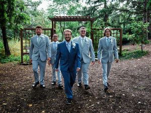 A wedding party in coordinating blue and gray suits walks together on a garden path under a wooden pergola.