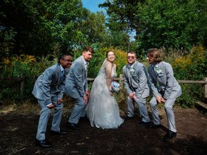 Wedding party in gray suits and white dress pose playfully in a garden setting on a sunny day.