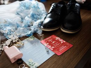 Wedding accessories including blue fabric, red invitation cards, and black shoes arranged on dark wooden surface.