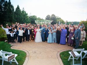 Large wedding party and guests gathered outdoors for a panoramic group photo on a sunny day.