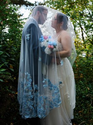 A wedding couple embraces under a delicate lace veil with blue floral embroidery in a garden setting.