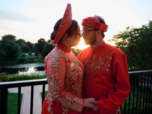 A couple in traditional red wedding attire standing on a deck at sunset near a lake.