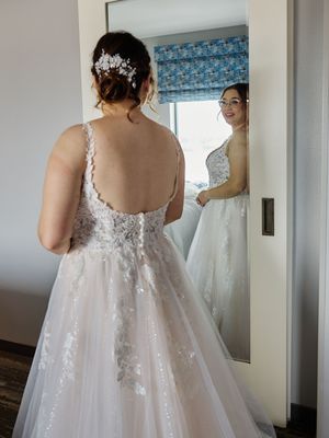A wedding dress with delicate lace details and a flowing white train is shown from behind in a dressing room.