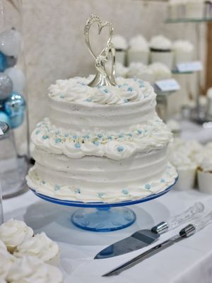 White two-tier wedding cake with blue accents and silver heart topper on a blue pedestal stand.