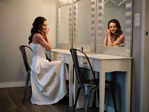 A bride in a white gown sits at a makeup vanity with mirrors and lights.