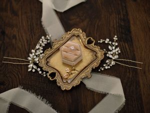 Wedding rings displayed on a vintage gold frame with pearl accents and white ribbon on a wooden surface.