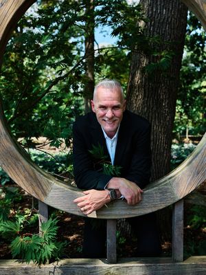 A person in a black jacket sits casually on a curved wooden bench surrounded by lush green trees in a garden setting.