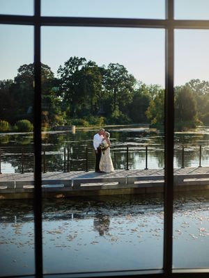 A couple stands on a wooden dock overlooking a serene lake with lily pads during golden hour.