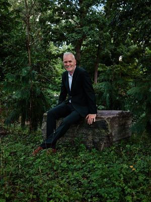 A person in a dark suit sits casually on a stone wall surrounded by lush green foliage in a wooded outdoor setting.
