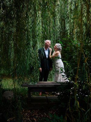 A couple sits together on a wooden bench under cascading willow tree branches in a romantic garden setting.