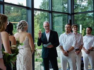 Wedding guests gather in an elegant room with floor-to-ceiling windows overlooking a scenic outdoor landscape.