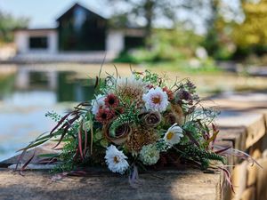 A rustic floral arrangement with white blooms and greenery sits on a wooden dock by a lake with buildings in the background.