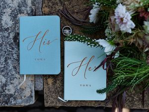 Two light blue wedding vow books with gold lettering labeled 'His' and 'Hers' displayed on stone with floral accents.