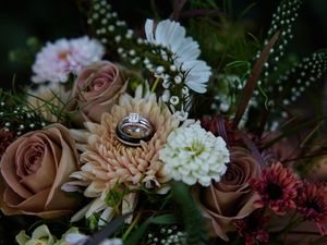 Wedding rings nestled among dusty pink roses, white daisies, and delicate baby's breath in a romantic floral arrangement.