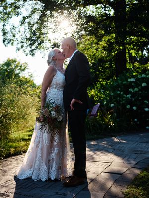 A couple in formal attire share an intimate moment on a stone patio surrounded by lush greenery in natural sunlight.