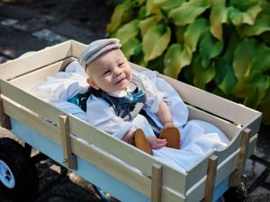 Young ring bearer in formal wear lying in rustic wooden wagon outdoors.