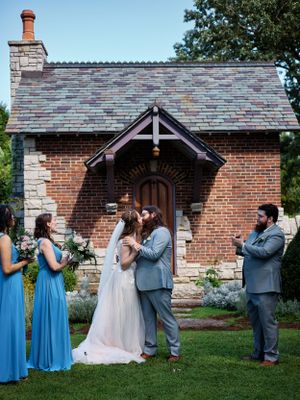 A wedding ceremony takes place in front of a charming brick chapel with blue-clad attendants.