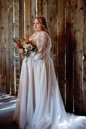 A bride in a lace wedding gown holds a beautiful bouquet while standing in a rustic barn setting.