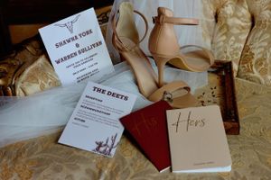 Wedding details flatlay with invitation, vows book, shoes and accessories on textured fabric.