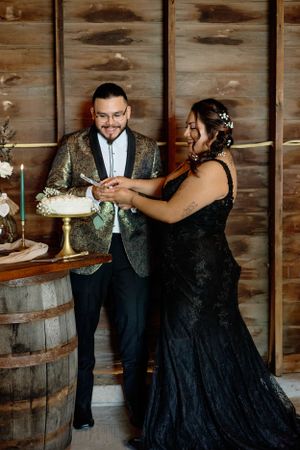 Couple cutting their wedding cake together in formal attire against rustic wooden backdrop.