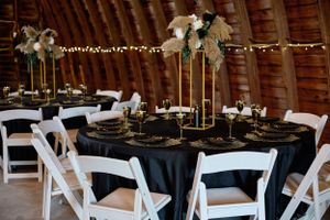 Elegant wedding reception table settings with black linens, gold accents and white folding chairs in a rustic barn venue.