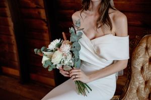 Bride in off-shoulder dress holds delicate white and green bouquet in rustic venue.
