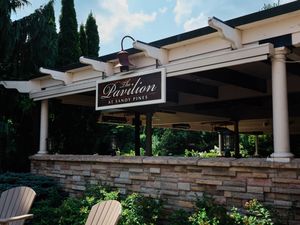 A modern outdoor pavilion with stone columns and white beams creates an elegant covered gathering space in a garden setting.