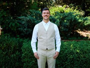 A person in a light beige vest and white shirt poses outdoors in a sunny garden setting with lush green foliage.