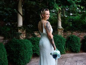 Bride in white lace dress holding blue bouquet poses in garden setting with brick wall and greenery.